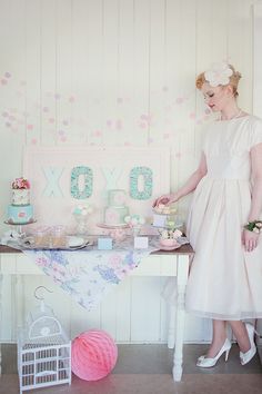 a woman in a white dress standing next to a table filled with cakes and cupcakes