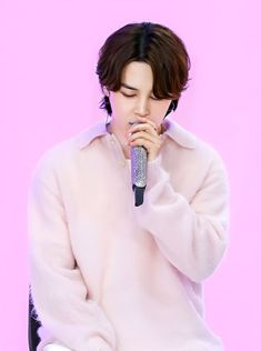 a male in a white sweater is holding a microphone and looking at the camera while standing against a pink background