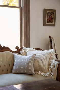 a couch with some pillows on top of it next to a table and chair in front of a window