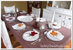 a dining room table set with white plates and silverware, fall leaves on the napkins