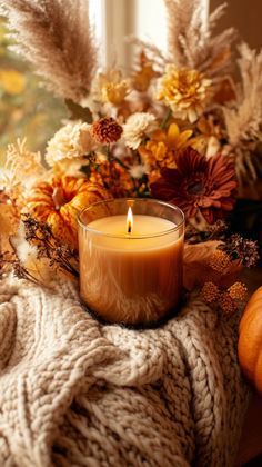 a candle sitting on top of a blanket next to flowers and pumpkins in front of a window