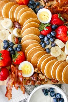 an assortment of breakfast foods including pancakes, eggs, fruit and bacon on a cutting board