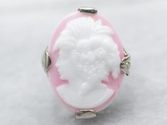 Sweet and full of detail, this Art Deco cameo ring is something special! The filigree is impressive, created using the die-stamping method that produces very fine detail while remaining strong and resilient. The pink glass cameo depicts a woman with 1920s styling, flowers set in curls, and an open neckline. Metal: 14K White Gold Gem: Glass Cameo Gem Measurements: 13.1 x 18.0 mm, Oval Ring Size: 3.25 SKU #: A36203 Each piece has been identified and graded by a Graduate Gemologist who has been cer Victorian Cabochon Wedding Rings, Round Intaglio Jewelry, White Oval Cameo Jewelry, Intaglio White Gold Ring, White Oval Rings With Intaglio, Pink Wedding Rings With Polished Finish, White Cameo Rings For Wedding, Luxury Cameo Ring Jewelry, Elegant White Cameo Ring