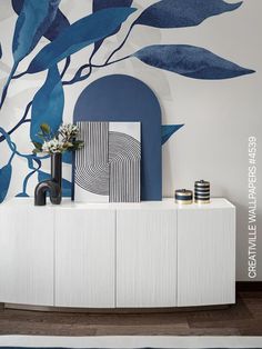 a white cabinet with blue leaves painted on the wall next to a painting and vase