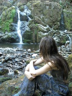 a woman sitting in front of a waterfall with her hands on her knees looking at the water