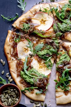 a pizza topped with apples, broccoli and other toppings next to a bowl of seeds