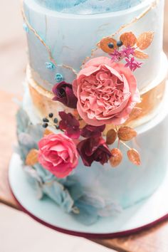 a three tiered blue cake with pink flowers on the top and gold trimmings