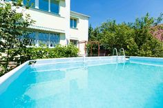 an empty swimming pool in front of a white house with trees and bushes around it