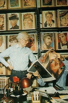 a man standing next to a desk with pictures on the wall behind him and a lamp in front of him