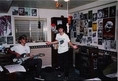 two young men are playing guitars in a room with posters on the wall behind them