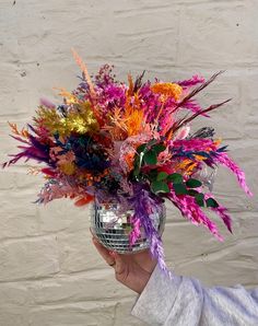 a person holding a disco ball filled with colorful flowers and feathers in front of a white brick wall