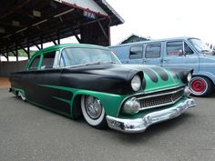 an old green and black car parked next to a blue van in a parking lot