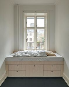 a bed sitting under a window in a bedroom next to a wooden shelf with drawers