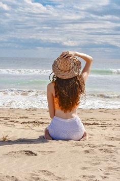 a woman sitting on top of a sandy beach next to the ocean wearing a hat