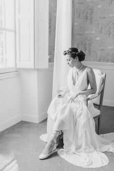 a woman in a white dress sitting on a chair next to a window with curtains