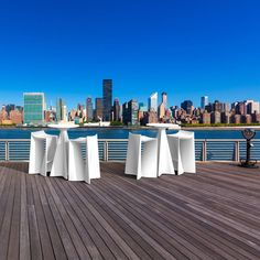 white tables and chairs on a wooden deck near the water with city skyline in background