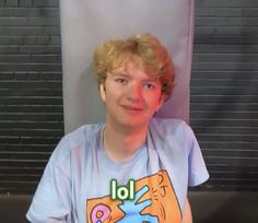 a young man with blonde hair wearing a blue t - shirt and looking at the camera