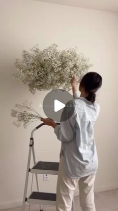 a woman standing on a stepladder painting a white wall with flowers in it