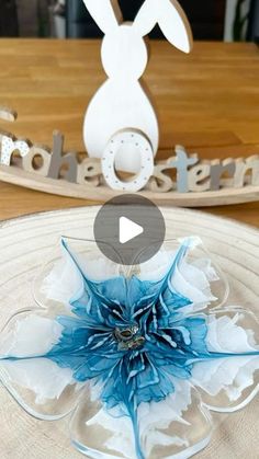 a white and blue paper flower sitting on top of a glass plate next to a wooden sign