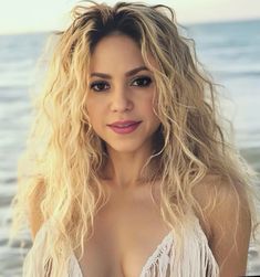 a woman with long blonde hair standing in front of the ocean wearing a white dress