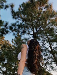 a woman with long hair standing in front of pine trees looking up at the sky