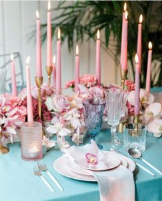 a blue table cloth with pink flowers and candles on it is set for a formal dinner