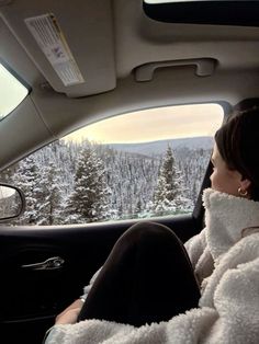 a woman sitting in the passenger seat of a car looking out at snow covered trees