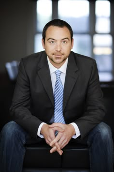 a man in a suit and tie sitting on a black chair with his hands crossed