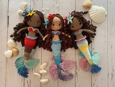 three crocheted mermaid dolls laying next to shells and seashells on a white wooden surface