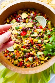 a person holding a tortilla chip in a wooden bowl filled with corn, avocado and salsa