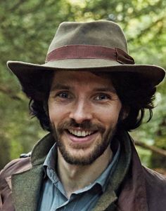 a man wearing a hat and jacket in the woods smiling at the camera with trees behind him