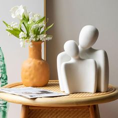 two white ceramic people sitting on a table next to a vase with flowers