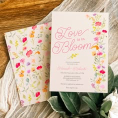 two wedding cards with flowers on them sitting next to some greenery and white flowers