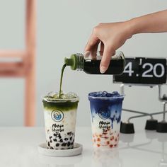 a person pouring green tea into two cups on top of a white table next to a wooden chair