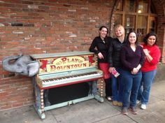 three women standing next to a piano on the side of a street in front of a brick building