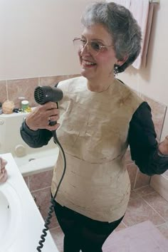 an older woman holding a hair dryer in her hand while standing next to a bathroom sink