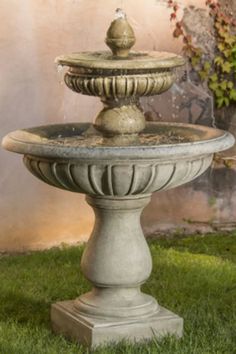 an outdoor fountain in the grass near a building with shutters on it's windowsill