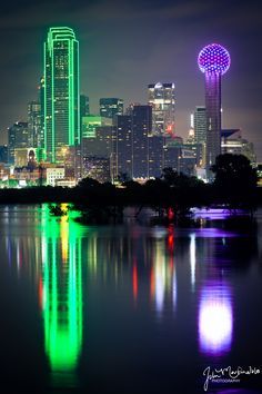 the city skyline is lit up in green, purple and blue colors with lights reflecting on the water