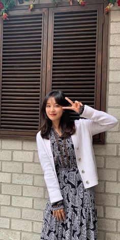 a woman standing in front of a brick wall with her hand on her head and wearing a white cardigan