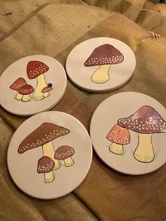 four coasters with mushrooms painted on them sitting on a tablecloth covered surface in front of a pillow