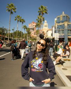 a woman in mickey mouse ears is standing on the street with her hands behind her back