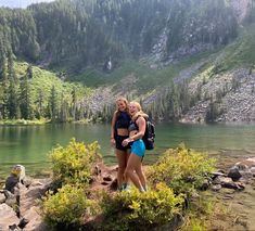 two women standing next to each other in front of a lake with mountains behind them