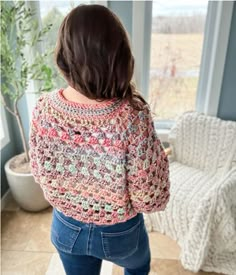 a woman standing in front of a window with her back to the camera, wearing jeans and a crocheted sweater