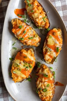 four baked potatoes with cheese and green onions on a white plate, sitting on a checkered table cloth