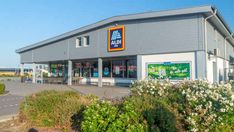 the exterior of a store with flowers in front and bushes around it, on a sunny day