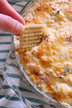 a person dipping a cracker into a cheesy dip in a glass dish