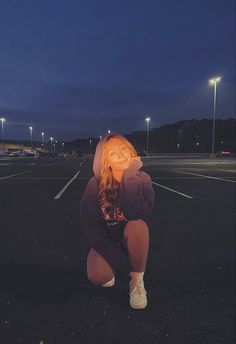 a woman kneeling down in the middle of an empty parking lot at night with lights on