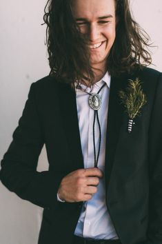 a man with long hair wearing a black suit and flower in his lapel is smiling at the camera