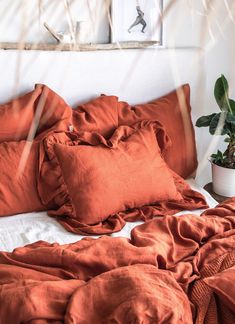 an unmade bed with orange sheets and pillows on it, next to a plant