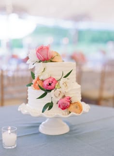 a white wedding cake with pink and orange flowers sitting on top of a table next to a candle